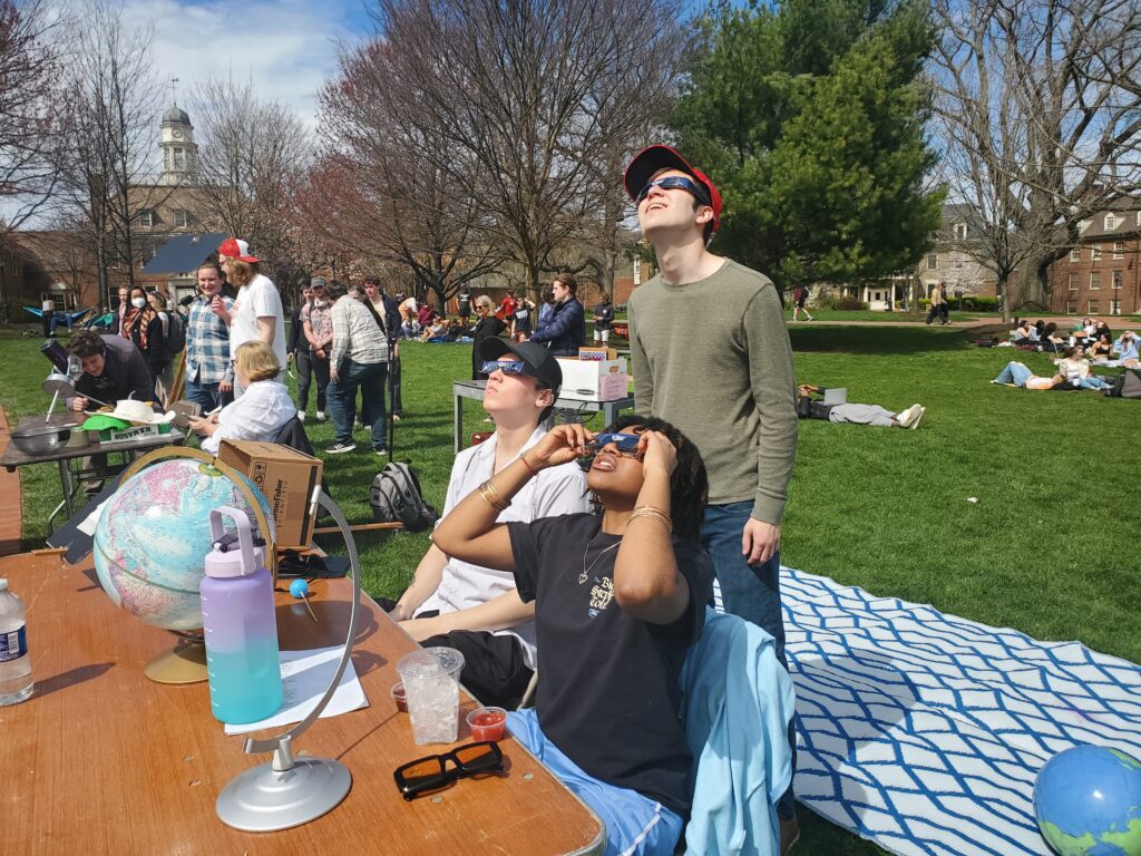Three college students behind a table gaze upwards wearing eclipse glasses. There is a globe on the table. 