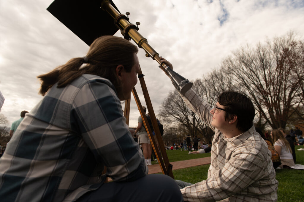 Two male college students view a sun funnel. One student has a long blond ponytail and blue shirt; he watches as a dark-haired student in a light shirt looks up at the telescope. The telescope in the background is a long brass refractor with a black plastic funnel attached to the bottom end. At the top/front end, a black board shields the viewer's eye from the sun. 