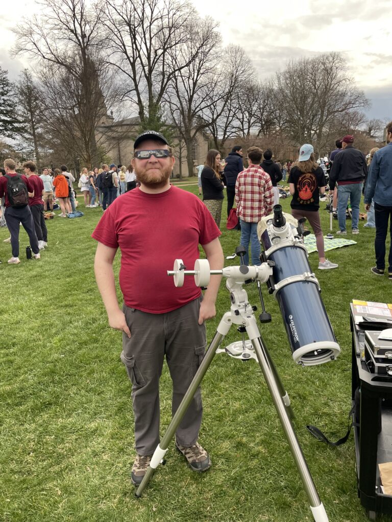 A male college student in a red shirt and baseball cap stands next to a blue Newtonian telescope on a white and silver tripod mount. The telescope has masking tape around the front, holding a solar filter on (the filter itself is not visible from this angle)